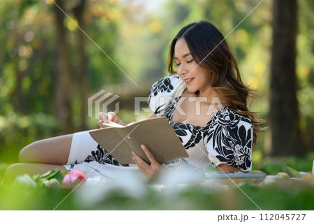Smiling millennial woman sitting on picnic blanket and reading book 112045727