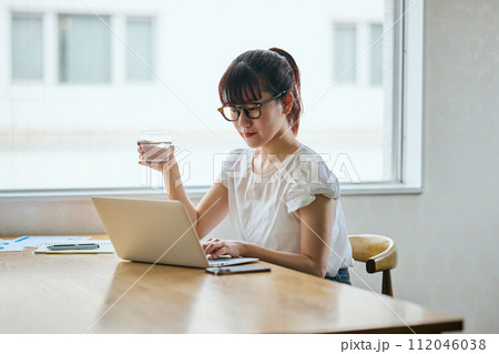 グラスを持ちながらパソコンをタイピングする若い女性 112046038