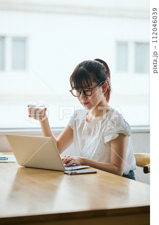 グラスを持ちながらパソコンをタイピングする若い女性 112046039