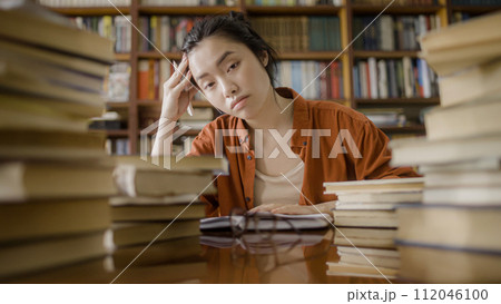 Portrait of exhausted asian woman student sitting behind stacks of books in a library 112046100