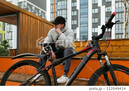 A man in a cap and a warm jacket enjoys a walk with a bicycle in the city A man in a cap and a warm jacket enjoys a walk with a bicycle in the city 112049414