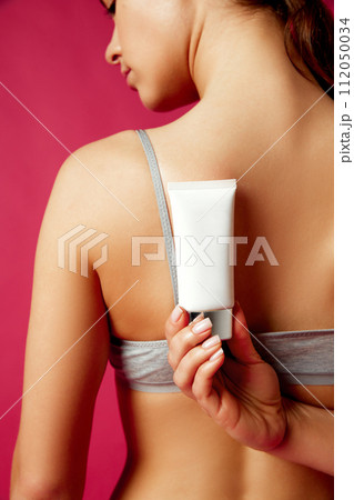 Woman holding cosmetic skincare tube near her shoulder against vivid pink studio background. 112050034