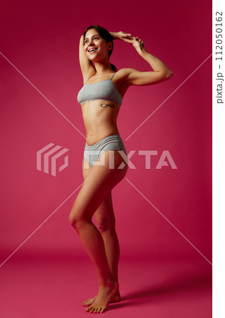 Full-length portrait of smiling woman posing in matching athletic wear holding hands behind her head against vivid pink studio background. 112050162