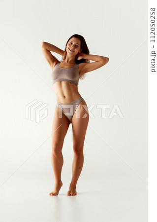 Full-length portrait of smiling woman posing in matching athletic wear holding hands behind her head against grey studio background. Full-length portrait of smiling woman posing in matching athletic wear holding hands behind her head against grey studio background. 112050258