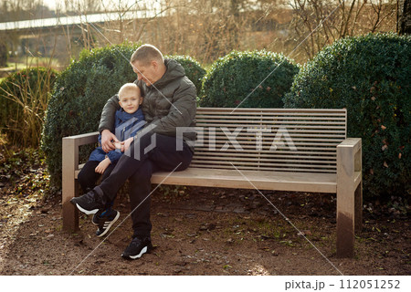 Autumnal Family Affection: Father, 40 Years Old, and Son - Beautiful 8-Year-Old Boy, Seated in the Park. 112051252