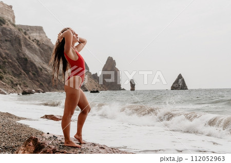 Woman summer travel sea. Happy tourist in red bikini enjoy taking picture outdoors for memories. Woman traveler posing on beach at sea surrounded by volcanic mountains, sharing travel adventure joy 112052963