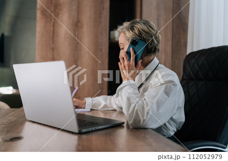 Elegant busy middle-aged business woman multitasking in office, working online with laptop, talking mobile phone, writing documents with pen sitting at office desk. Female employee talking on phone. Elegant busy middle-aged business woman multitasking in office, working online with laptop, talking mobile phone, writing documents with pen sitting at office desk. Female employee talking on phone. 112052979