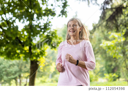 Active senior woman with a smile jogging in a lush green park, embracing fitness and health in the golden years. 112054398