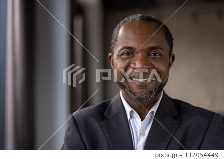 Portrait of a positive African American businessman with a warm smile, dressed in a formal suit, at a workplace. 112054449