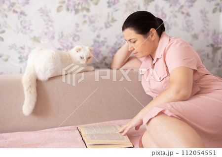 Woman in pink dress reading a book with a white Scottish Fold resting on the couch. Woman in pink dress reading a book with a white Scottish Fold resting on the couch. 112054551