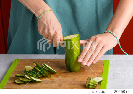 Woman's hands cutting zucchini on wooden cutting board at home, preparing healthy food, close up Woman's hands cutting zucchini on wooden cutting board at home, preparing healthy food, close up 112056059