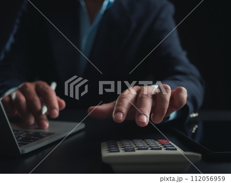 Businessman in black suit working on laptop computer and tablet and calculator, Hand Hands touch on tablet at office with dark background, Online working, Close up, Copy space. 112056959