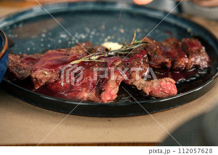 Man Cutting Rib Eye Steak, Pouring it Red Sauce on a Black Plate. Person Having a Delicious Lunch. Grilled Sirloin Beef Steak with Rosemary and Garlic on a Wooden Table. BBQ Meat Delicacy Bio Food. Man Cutting Rib Eye Steak, Pouring it Red Sauce on a Black Plate. Person Having a Delicious Lunch. Grilled Sirloin Beef Steak with Rosemary and Garlic on a Wooden Table. BBQ Meat Delicacy Bio Food. 112057628
