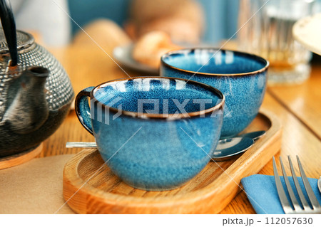 Brewing and Serving Tea. Teapot Pouring Green Tea in Blue Ceramic Cup on Wooden Cafe Table. Two Pottery Handmade Mugs Early in the Morning. Trendy Authentic Porcelain Set. Breakfast Concept. Tea Time 112057630