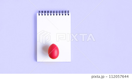 Notebook with red Easter Egg. Spiral notepad on spring violet background. Top view of open writing pad with blank page, office flat lay. White Page for your write. Copy Space. Easter Mock Up 112057644