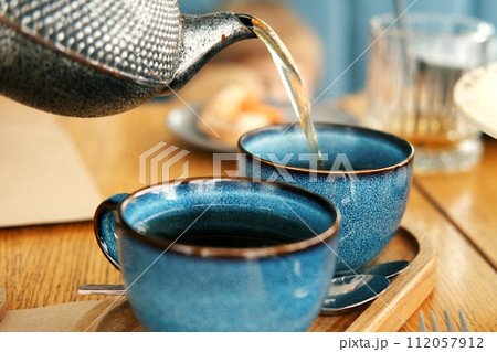 Brewing and Serving Tea. Teapot Pouring Green Tea in Blue Ceramic Cup on Wooden Cafe Table. Two Pottery Handmade Mugs Early in the Morning. Trendy Authentic Porcelain Set. Breakfast Concept. Tea Time 112057912