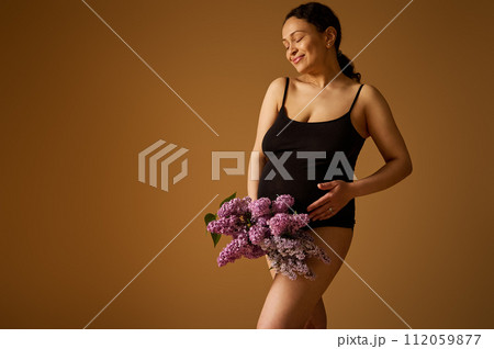 Pregnant woman with big belly in last pregnancy trimester, in black lingerie, holding lilacs, isolated beige backdrop 112059877