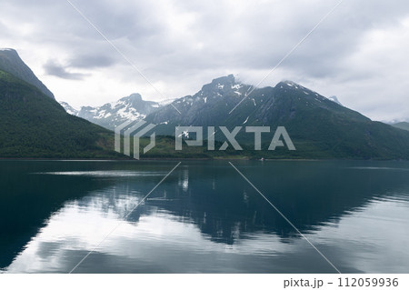 The stillness of a Norwegian fjord reflects cloudy skies and shadowy mountains, a moody landscape 112059936
