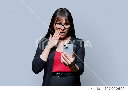 Surprised middle-aged woman looking at smartphone on gray background 112060602