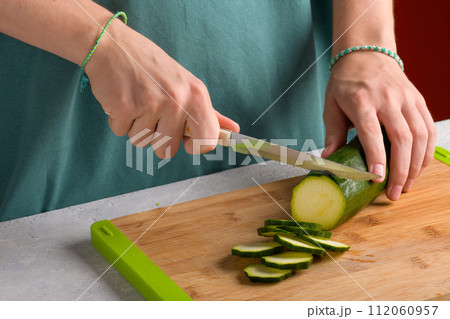 Woman's hands cutting zucchini on wooden cutting board at home, preparing healthy food, close up Woman's hands cutting zucchini on wooden cutting board at home, preparing healthy food, close up 112060957