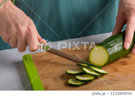 Woman's hands cutting zucchini on wooden cutting board at home, preparing healthy food, close up Woman's hands cutting zucchini on wooden cutting board at home, preparing healthy food, close up 112060958