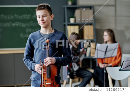 Medium portrait of confident teen boy holding violin standing in classroom looking at camera, his music teacher and classmate sitting in background 112062174