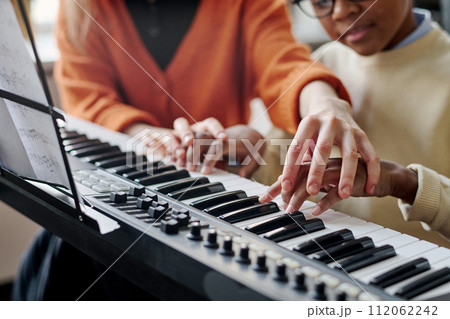 Selective focus shot of unrecognizable woman teaching Black boy to play synthesizer at music class 112062242