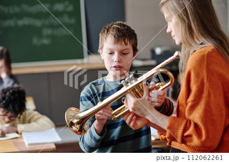Selective focus medium shot of young female teacher giving trumpet to boy during music class 112062261