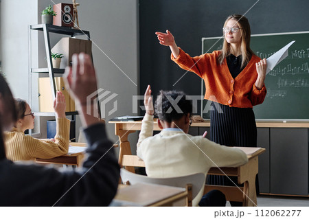 Young Caucasian female music teacher asking question, her students sitting at desks raising hands to answer it Young Caucasian female music teacher asking question, her students sitting at desks raising hands to answer it 112062277
