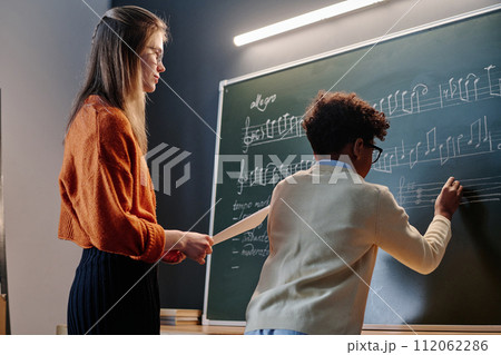 Medium shot of young Caucasian female music teacher watching her Black student writing notes on blackboard Medium shot of young Caucasian female music teacher watching her Black student writing notes on blackboard 112062286