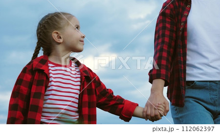 Girl ambles through corn field hand in hand with farmer mom. Daughter has weekend with mother guided by hand through corn field. Girl and agriculturist spend time together among corn field 112062397