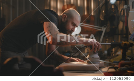 Male craftsman grinds wooden guitar body using grinding machine Male craftsman grinds wooden guitar body using grinding machine 112063090
