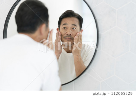 鏡を見ながら頬に手をあてる笑顔の50代半ばの男性 112065529