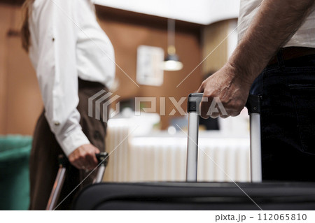 Close view of couple entering foyer of fancy hotel with trolley bags. Detailed shot of elderly individuals grasping suitcases as they walk up to registration desk to check in with the receptionists. Close view of couple entering foyer of fancy hotel with trolley bags. Detailed shot of elderly individuals grasping suitcases as they walk up to registration desk to check in with the receptionists. 112065810
