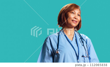 Happy proficient BIPOC nurse smiling while looking at camera. Portrait of cheerful asian healthcare specialist wearing professional scrubs, isolated over blue studio background Happy proficient BIPOC nurse smiling while looking at camera. Portrait of cheerful asian healthcare specialist wearing professional scrubs, isolated over blue studio background 112065838