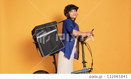 BIPOC courier woman taking break between takeout meal deliveries, texting friends. Cyclist waiting to receive next customer food order, isolated over orange studio background 112065841