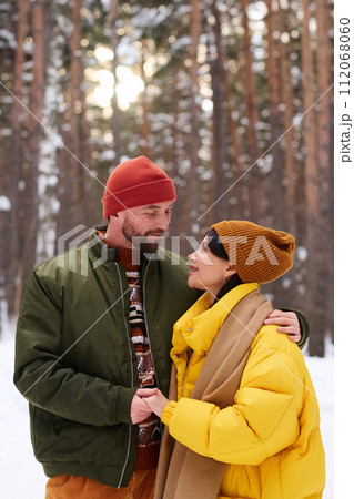Vertical medium portrait of married couple wearing casual warm clothes smiling to each other while spending time in forest on winter day Vertical medium portrait of married couple wearing casual warm clothes smiling to each other while spending time in forest on winter day 112068060