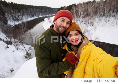 Cheerful Caucasian man and woman in love standing on cliff against river taking selfie on smartphone 112068130