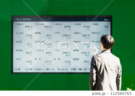 株価ボードの電光掲示板で銘柄を見る30代のビジネスマン投資家男性の後ろ姿 株価ボードの電光掲示板で銘柄を見る30代のビジネスマン投資家男性の後ろ姿 112068763