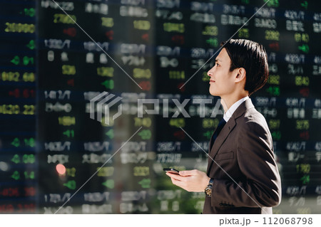 NISAでの運用を考え株価ボードの前でスマートフォンのアプリで株取引をする30代の男性 NISAでの運用を考え株価ボードの前でスマートフォンのアプリで株取引をする30代の男性 112068798