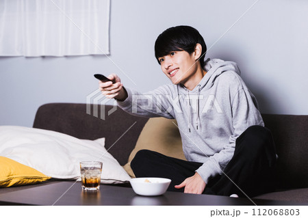部屋でお酒とツマミを肴にテレビドラマやアニメを楽しむ為にリモコンを操作する30代の男性 部屋でお酒とツマミを肴にテレビドラマやアニメを楽しむ為にリモコンを操作する30代の男性 112068803