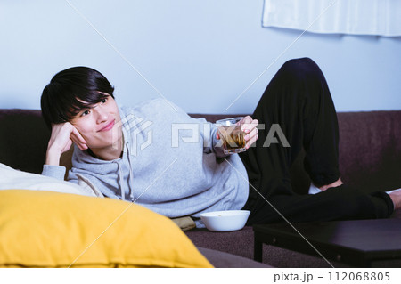 ソファに寝っ転がってお酒を飲みながらリラックスしてTVやサブスクリプションコンテンツを鑑賞する男性 112068805