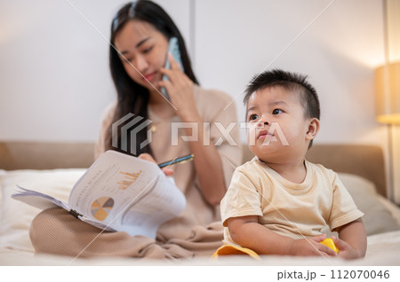 A cute Asian little boy is playing toys in bed while his mother is working and talking on the phone. A cute Asian little boy is playing toys in bed while his mother is working and talking on the phone. 112070046