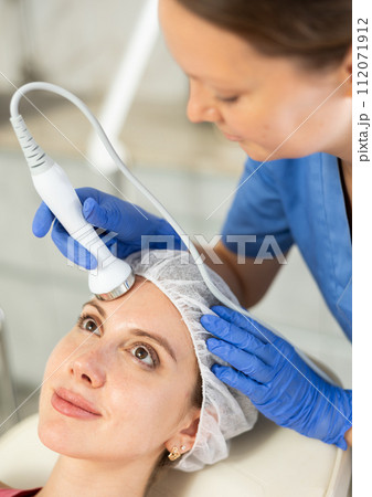 Young girl visitor receiving hardware electric massage and moisturizing bottom layer of skin by beautician in beauty clinic. Hardware cosmetology Young girl visitor receiving hardware electric massage and moisturizing bottom layer of skin by beautician in beauty clinic. Hardware cosmetology 112071912