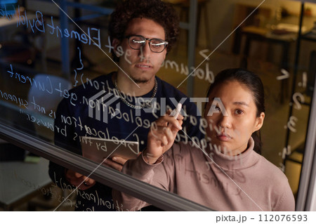 High angle of two young programmers working together on code writing it on glass wall in modern office 112076593