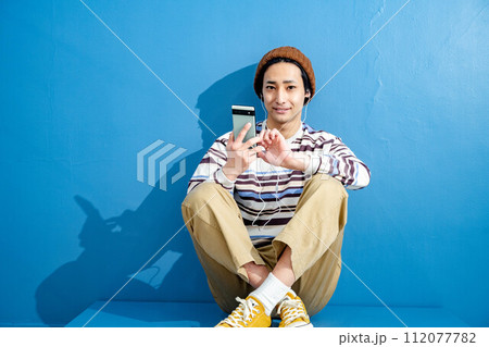 ブルーバックで帽子をかぶりスマートフォンを操作している若い男性 ブルーバックで帽子をかぶりスマートフォンを操作している若い男性 112077782