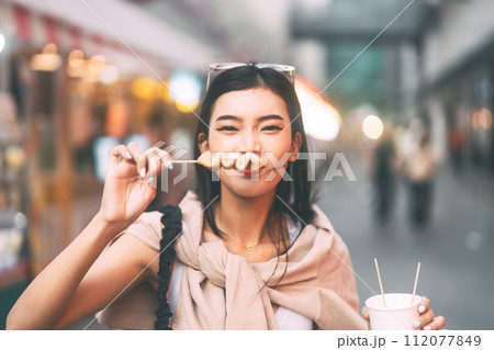 Happy asian foodie woman eating street food holding korea skewer oden Happy asian foodie woman eating street food holding korea skewer oden 112077849