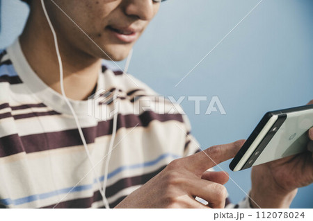 ブルーバックでメガネをかけて帽子をかぶりながらスマートフォンをいじっている若い男性 112078024