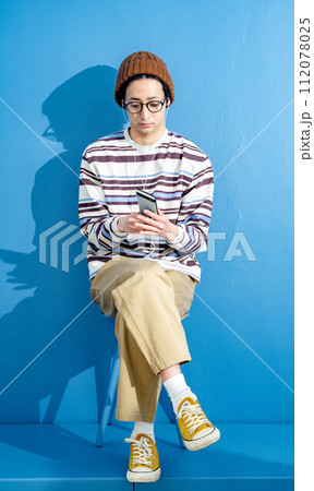 ブルーバックでメガネをかけて帽子をかぶりながらスマートフォンをいじっている若い男性 112078025