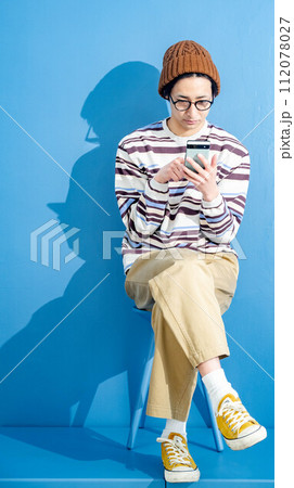 ブルーバックでメガネをかけて帽子をかぶりながらスマートフォンをいじっている若い男性 ブルーバックでメガネをかけて帽子をかぶりながらスマートフォンをいじっている若い男性 112078027
